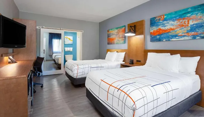 The image shows a modern and neatly arranged hotel room with twin beds colorful wall art and a separate space visible through sliding doors