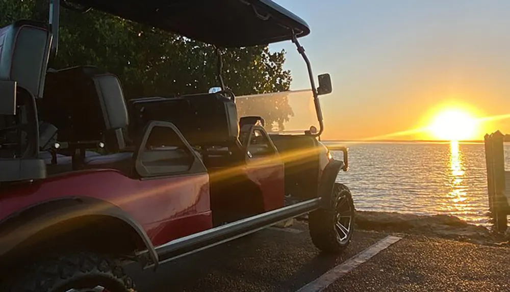 A golf cart is parked near a serene coastline at sunset with the sun casting a golden glow over the water