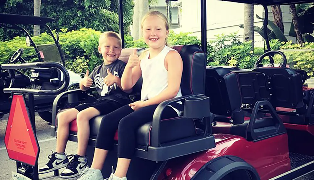 Two children are smiling and giving thumbs-up signs while sitting in a red golf cart