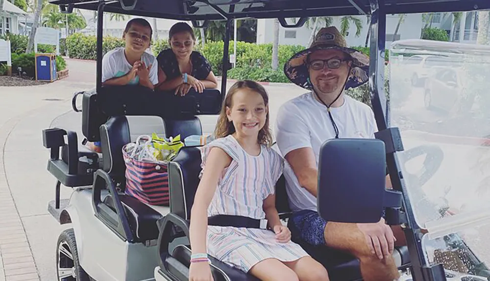 A group of four people likely a family is smiling for a photo while seated inside a golf cart with lush greenery in the background