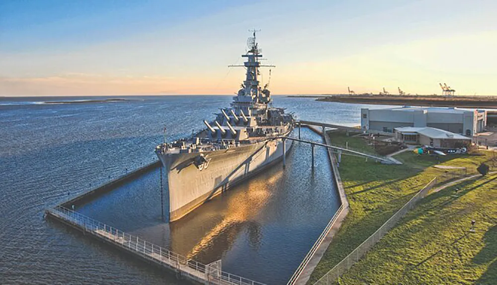 A retired battleship is docked as a museum next to a calm body of water under a clear sky at sunset