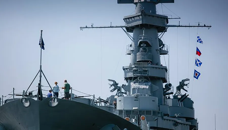 A group of individuals is standing on the deck of a naval ship adorned with signal flags, with some looking towards the camera.