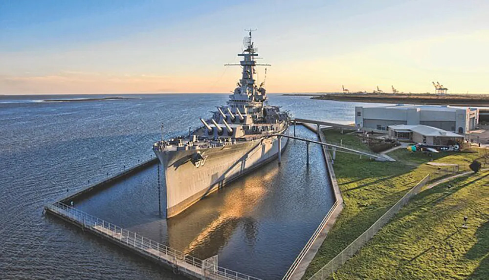 The image shows a large battleship moored in a water dock against a backdrop of open water and a clear sky during sunset or sunrise