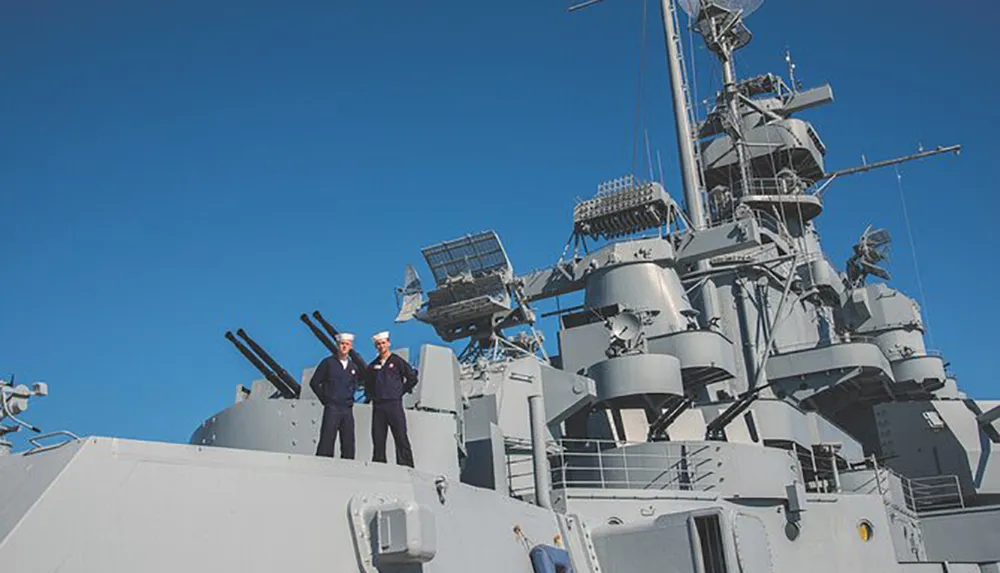 Two individuals are standing in naval attire on the deck of a naval ship with large gun turrets and radar equipment against a clear blue sky