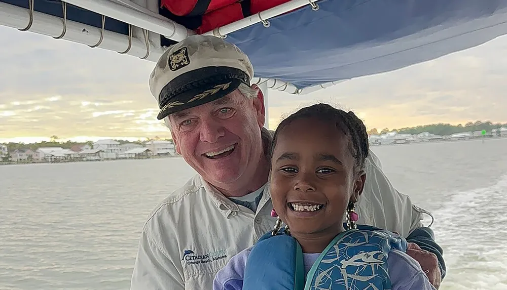An adult in a captains hat and a smiling child wearing a life jacket pose for a photo on a boat with waterfront houses in the background
