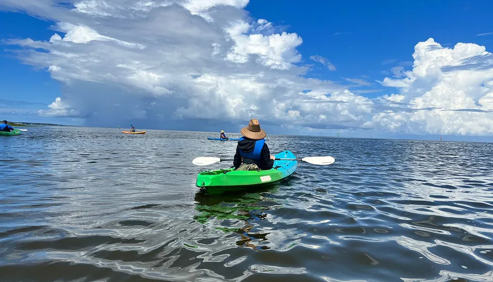 A person wearing a straw hat is kayaking on calm waters under a partly cloudy sky accompanied by other kayakers in the distance