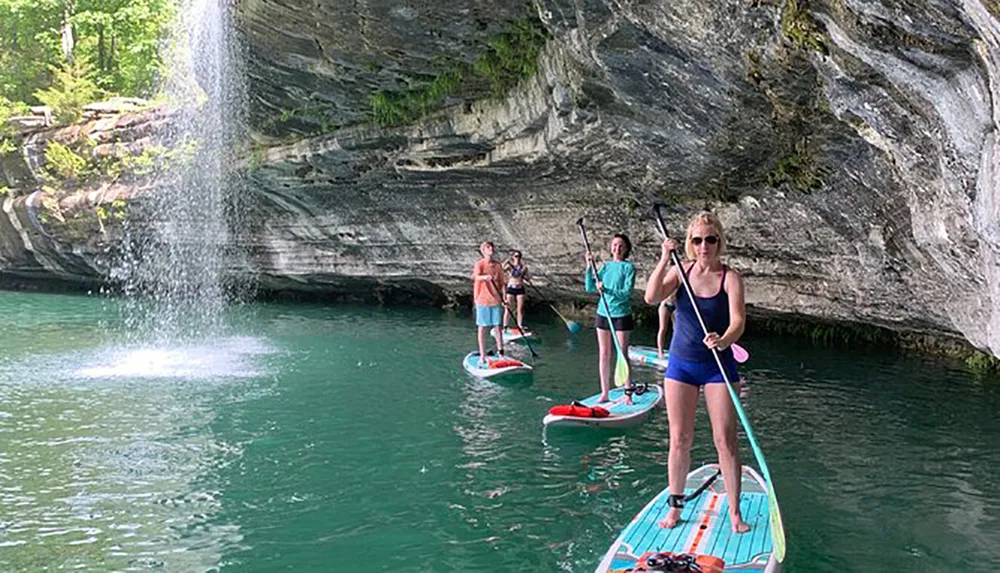 A group of people are paddleboarding near a waterfall and a rocky cliff