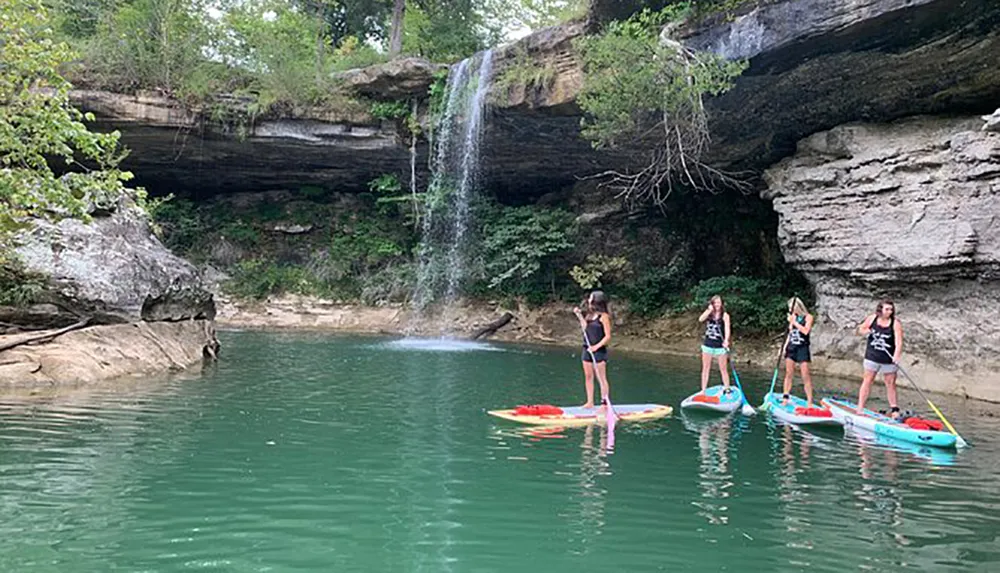 The image shows a group of people on paddleboards enjoying the serene setting of a waterfall cascading into a lush calm lagoon