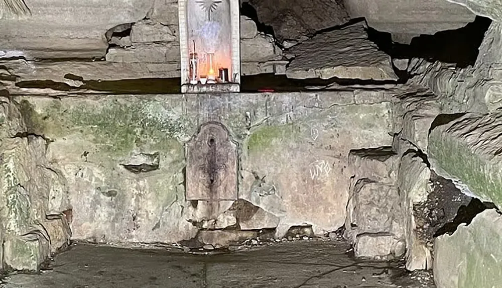 The image shows a small makeshift shrine or memorial located in a recess of a stone wall surrounded by rough rock surfaces possibly within a cave or under an overhang