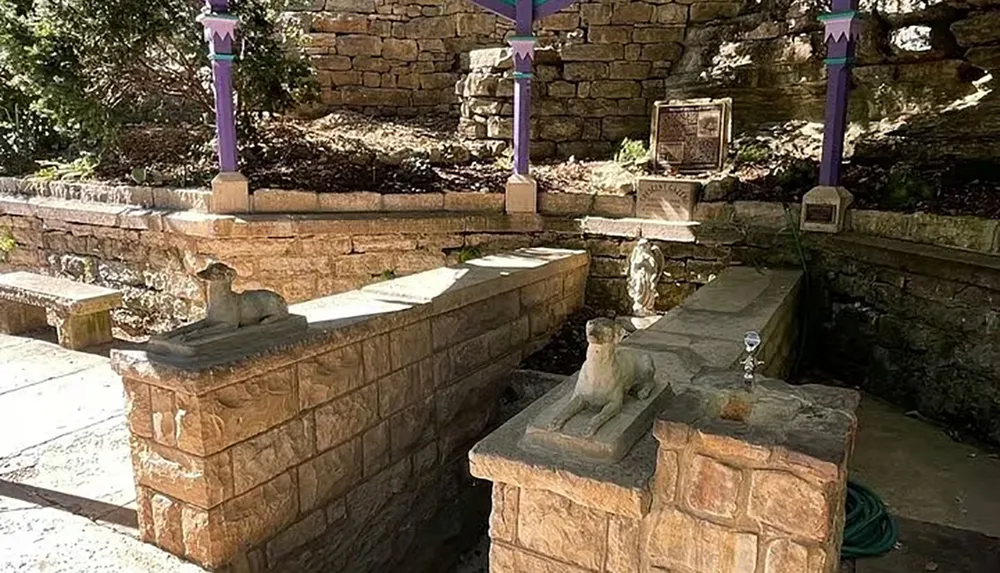 The image shows an outdoor memorial with inscribed stone benches a statue of a dog a hose lying on the ground and a purple pole set among a landscaped stone wall backdrop