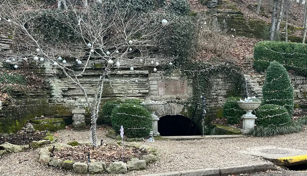 An old stone fountain with string lights stands before an arched entrance nestled within a weathered stone wall flanked by neatly trimmed hedges and bare trees evoking a sense of historical charm