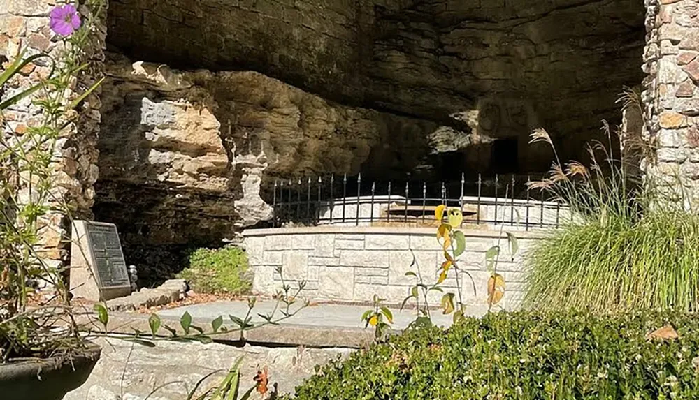 The image shows a stone structure with an arched opening leading into a cave-like space surrounded by greenery and a fence with a commemorative plaque visible on the left