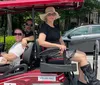 Three people are smiling and seated in a red golf cart parked on a city street on a sunny day