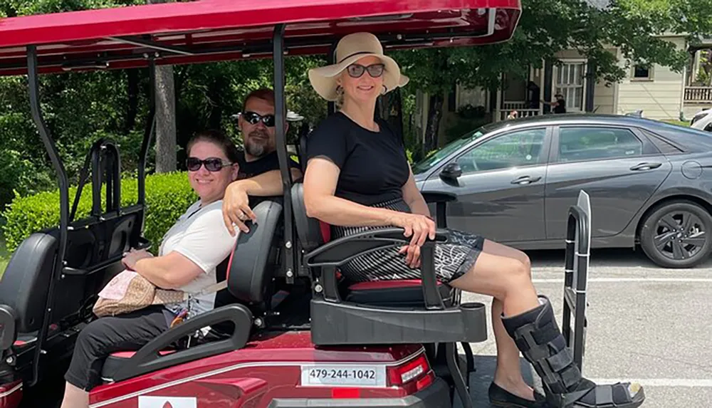 Three people are smiling and seated in a red golf cart parked on a city street on a sunny day