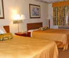 The image shows a standard hotel room with two double beds matching bedspreads framed artwork on the walls a window with curtains and a nightstand with a lamp and telephone