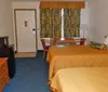 The image shows a standard hotel room with two double beds matching bedspreads framed artwork on the walls a window with curtains and a nightstand with a lamp and telephone