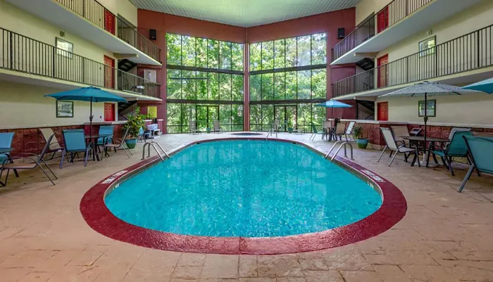 The image shows an indoor pool area with a blue swimming pool surrounded by seating arrangements enclosed by a building with multiple floors of balconies and a backdrop of large windows revealing trees outside