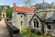 The image shows a historic stone building with arched windows and a domed roof, adjacent to a walkway bordered by a low stone wall, set within a lush, green landscape.