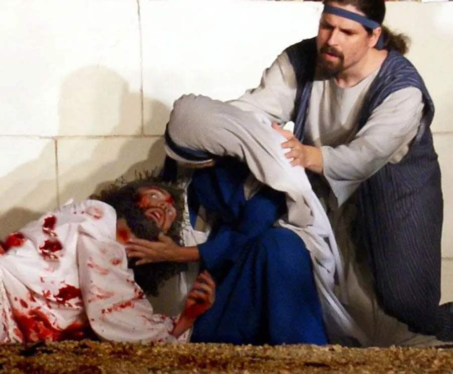 The image depicts a dramatic scene where two individuals dressed in historical garments appear to be tending to a third person who is covered in blood.