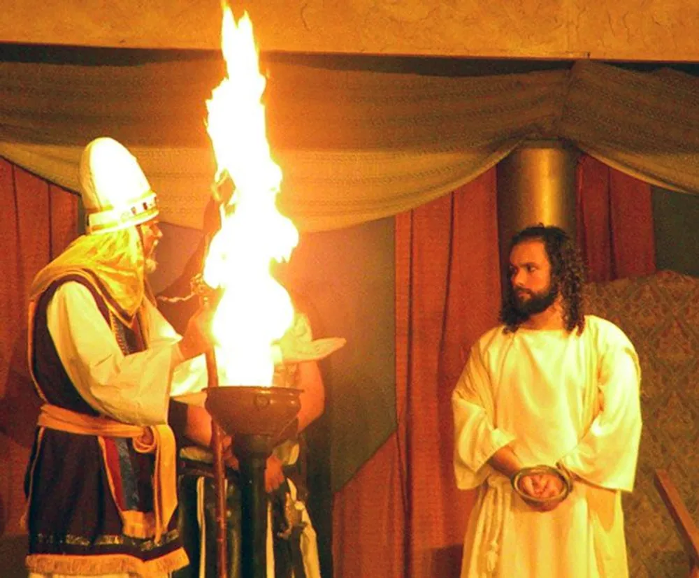 A person dressed in ancient robes stands by a large flame in a torch while another person in white watches intently