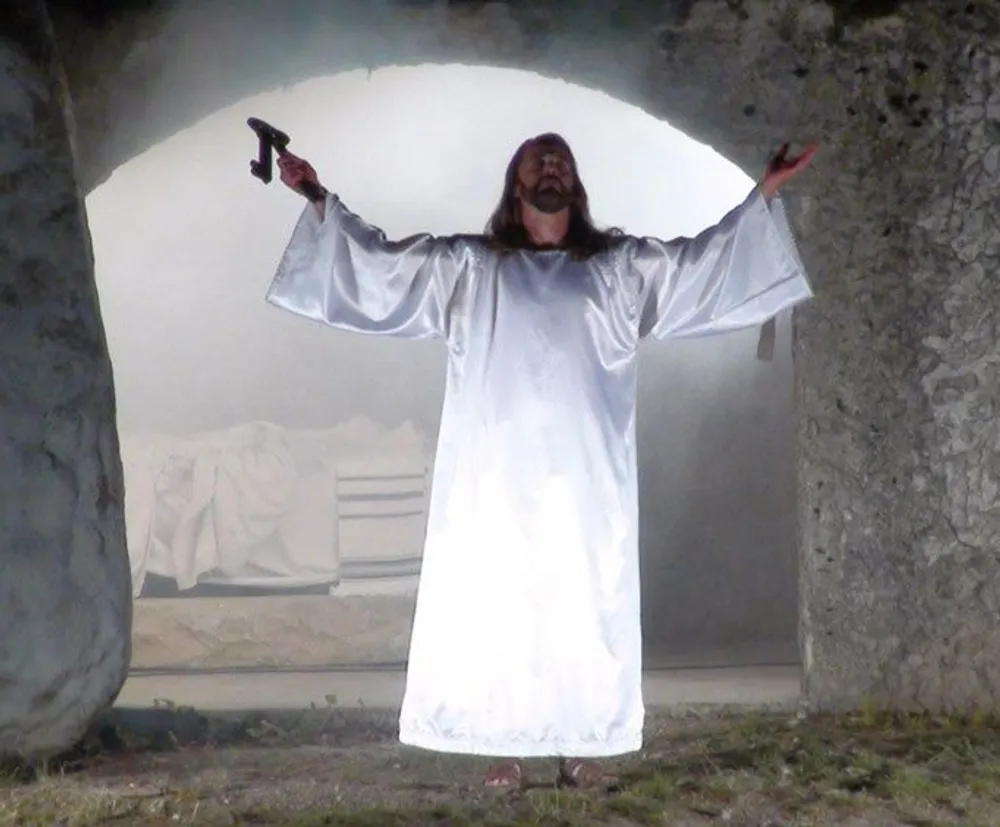 A person dressed in a white robe is standing with arms outstretched holding a small object in one hand framed by an arched stone entryway with a foggy backdrop