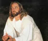 The image shows a person portraying a historical or religious figure traditionally identified with Jesus wearing a white robe and looking upwards with a contemplative expression