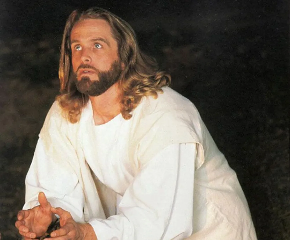 The image shows a person portraying a historical or religious figure traditionally identified with Jesus wearing a white robe and looking upwards with a contemplative expression