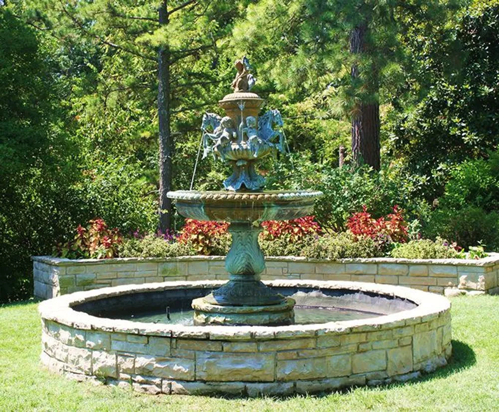 An ornate multi-tiered fountain is surrounded by a circle of flowers within a manicured garden setting