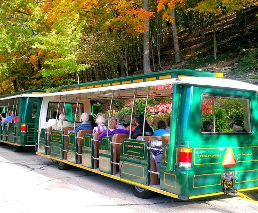 A tram full of passengers is touring through a wooded area with autumn foliage