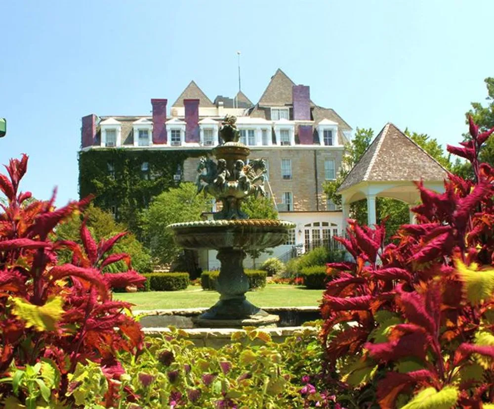 A stately building with a classic design overlooks a vibrant garden featuring a decorative fountain set under a clear blue sky