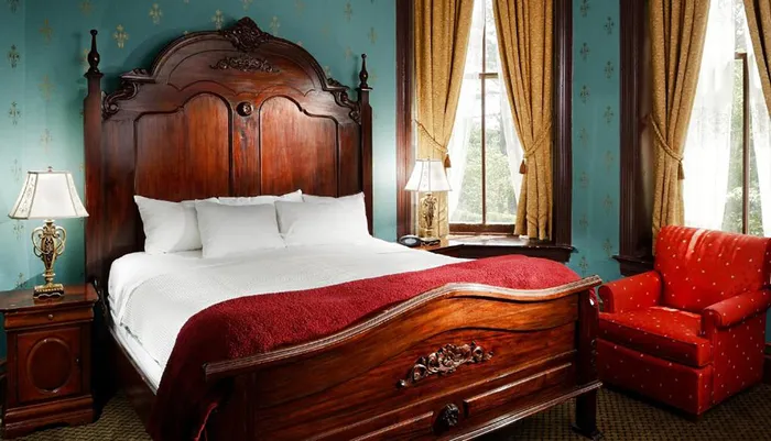 The image shows a classically decorated bedroom with an ornate wooden bed red bedding traditional furniture and heavy drapes framing a window