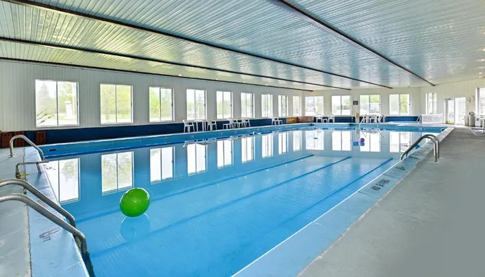The image shows an indoor swimming pool with a green ball floating on the water surrounded by chairs and large windows that offer a view of the outside