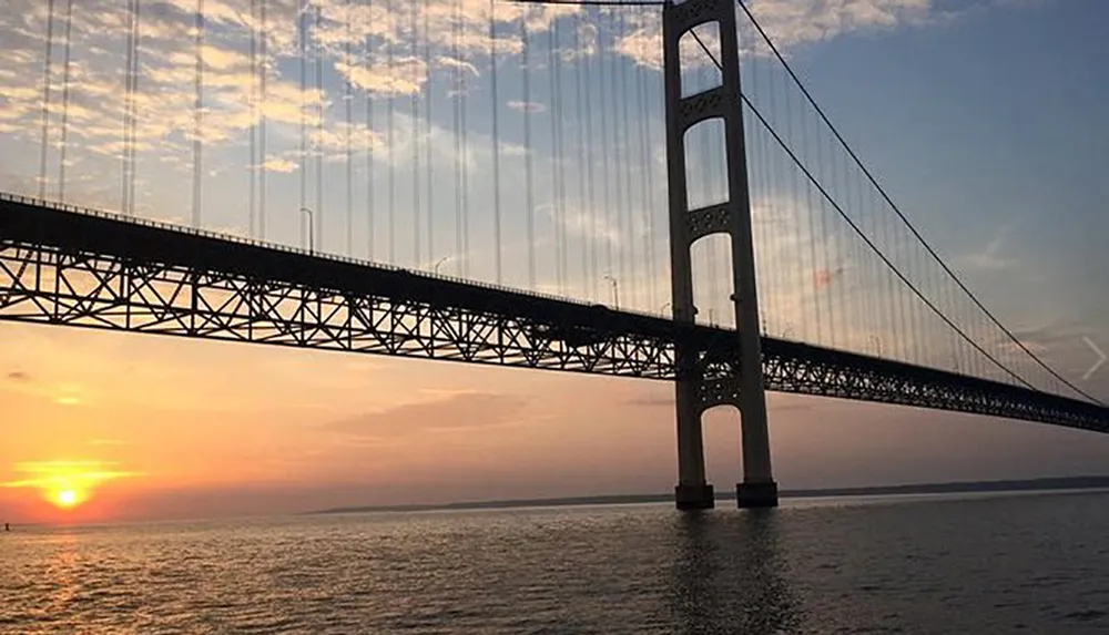 The image captures a majestic suspension bridge silhouetted against an evening sky with the sun setting over a calm body of water