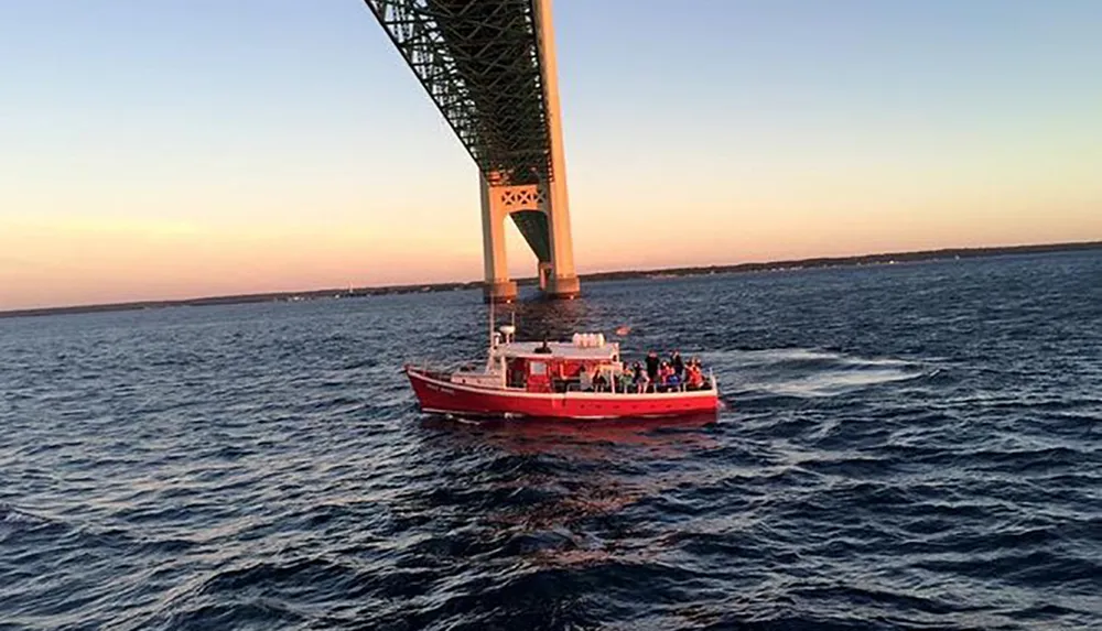 A red boat with passengers is sailing under a large bridge during the evening