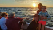 A group of people enjoys a scenic sunset aboard a boat, with a child in sunglasses smiling at the forefront.