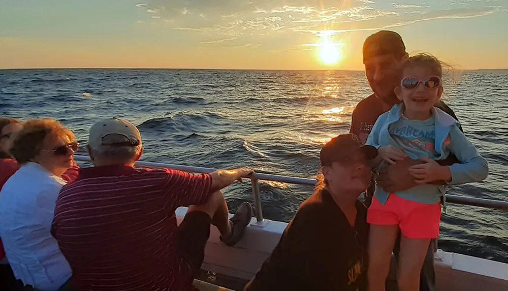 A group of people enjoys a scenic sunset aboard a boat with a child in sunglasses smiling at the forefront