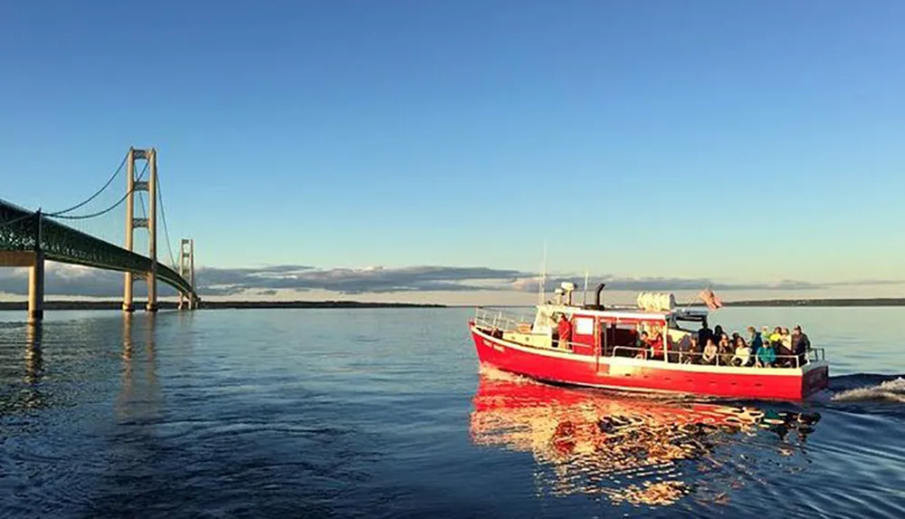 A red tour boat filled with passengers cruises on tranquil water near a large suspension bridge under a clear blue sky