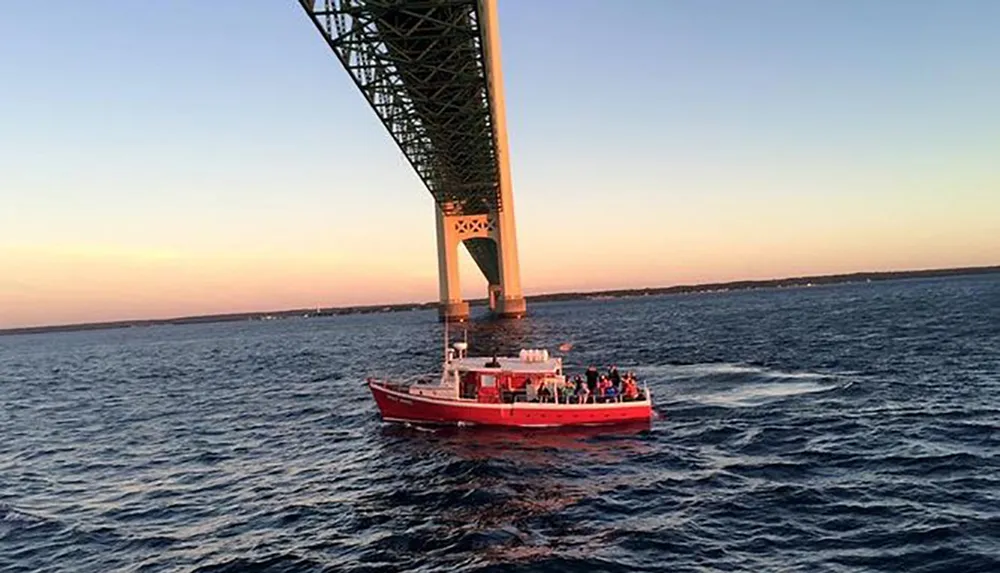 A red boat carrying passengers is sailing under a large bridge at dusk