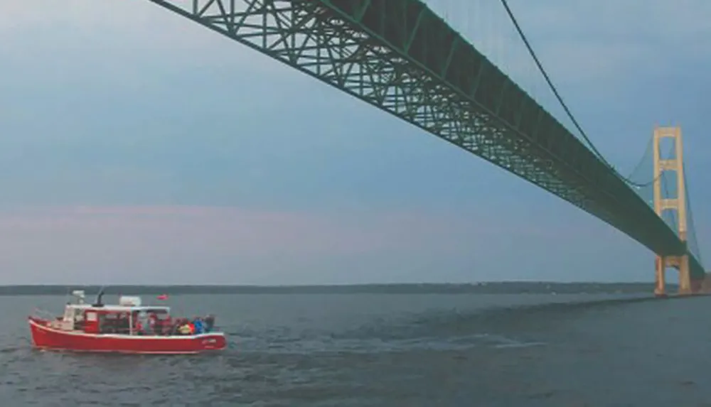 A red boat is passing under a large suspension bridge over a body of water