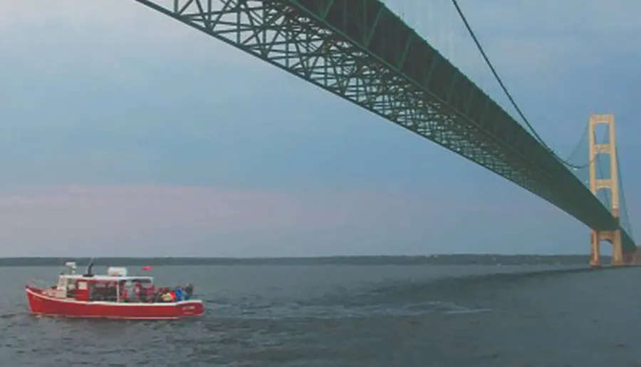 A red boat is passing under a large suspension bridge over a body of water.