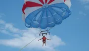 A person is parasailing under a blue and red parachute against a backdrop of a clear sky with a few clouds.