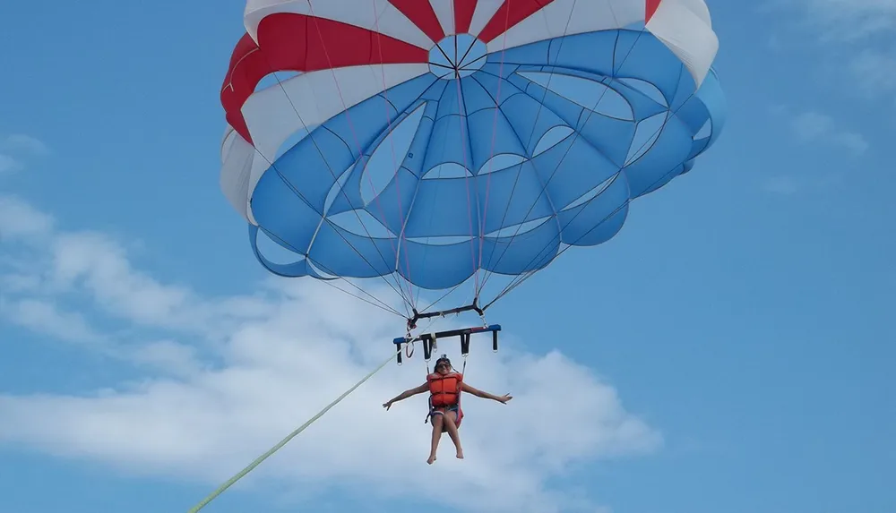 A person is parasailing under a blue and red parachute against a backdrop of a clear sky with a few clouds