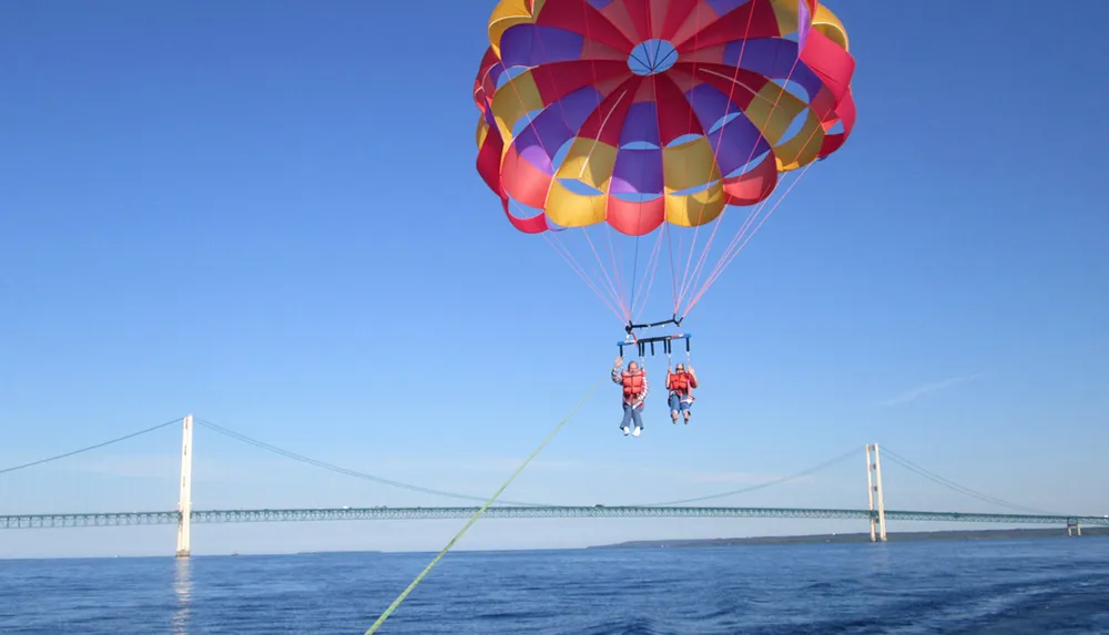 A colorful parachute carries two people in a tandem parasailing adventure over a calm blue sea with a long bridge in the background