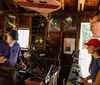 A blacksmith is working on a piece of metal with a hammer as two onlookers watch attentively in a rustic workshop setting