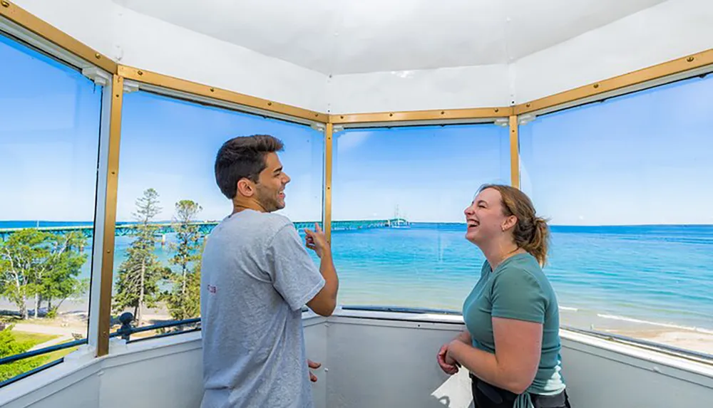 Two people are sharing a conversation and a cheerful moment inside a glass-enclosed structure with a picturesque view of the blue sea and sky