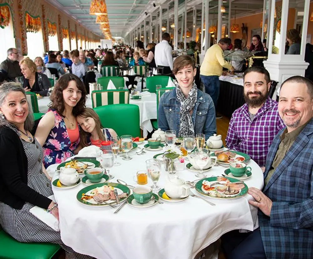 A group of people is smiling at the camera while sitting at a dining table with various dishes and beverages in a busy and ornately decorated restaurant