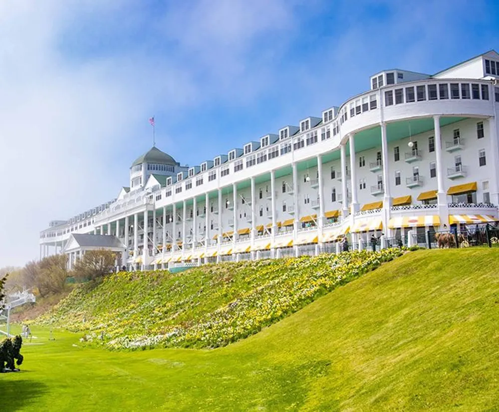 The image shows a grand multi-storied white hotel with a long porch lined with yellow awnings set against a clear blue sky with a vibrant green lawn and yellow flowers in the foreground