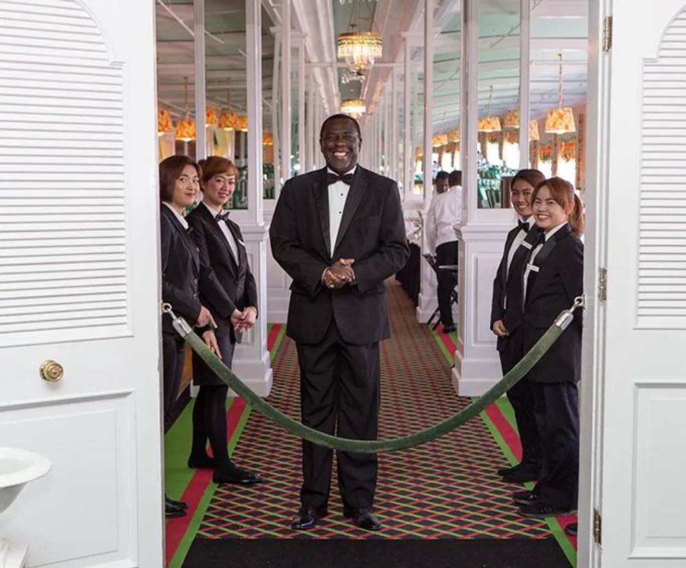 A smiling man in a suit stands at the entrance of an elegant corridor holding a rope barrier as several uniformed staff members look on