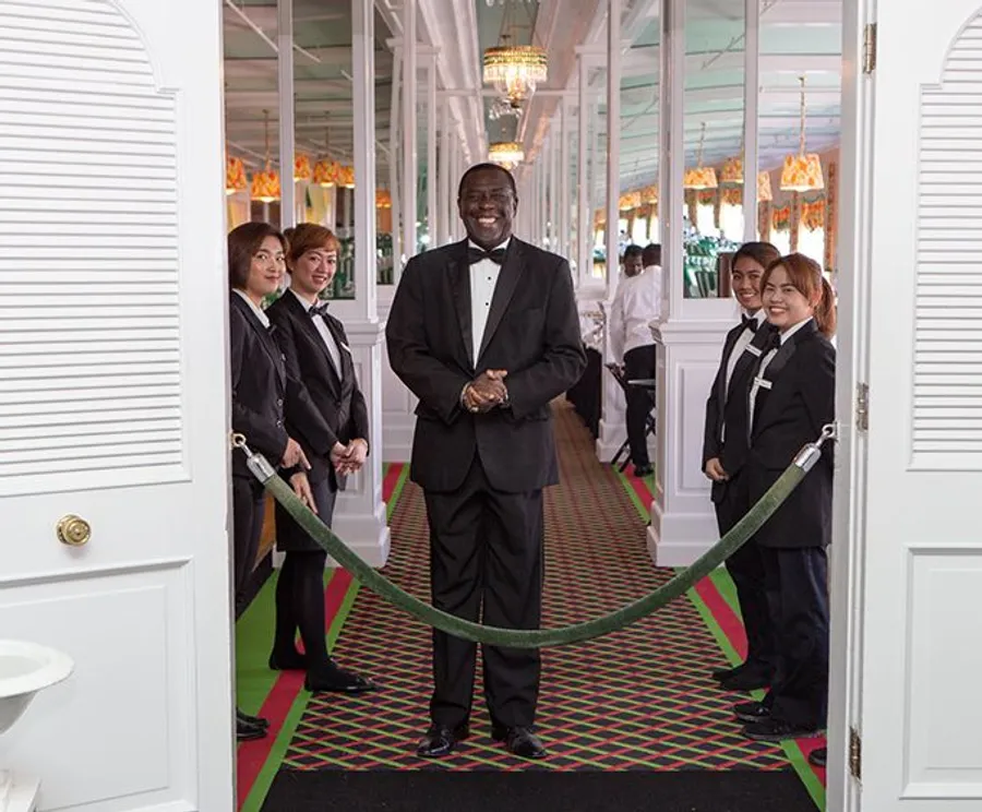 A smiling man in a suit stands at the entrance of an elegant corridor, holding a rope barrier, as several uniformed staff members look on.