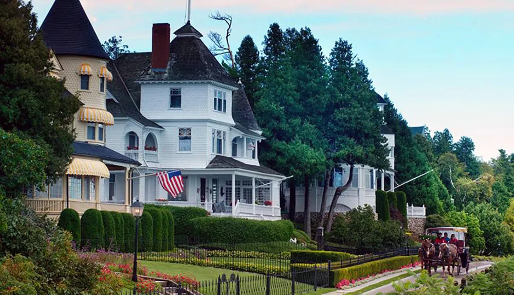 A horse-drawn carriage passes by a picturesque Victorian-style house adorned with American flags and surrounded by manicured gardens